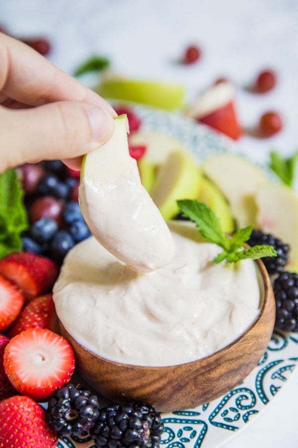 Small bowl of yogurt fruit dip on a platter that's surrounded by colorful fresh fruits including black berries, apples slices, strawberries, and blueberries. There's a hand holding an apple slice that has been dipped in the dip