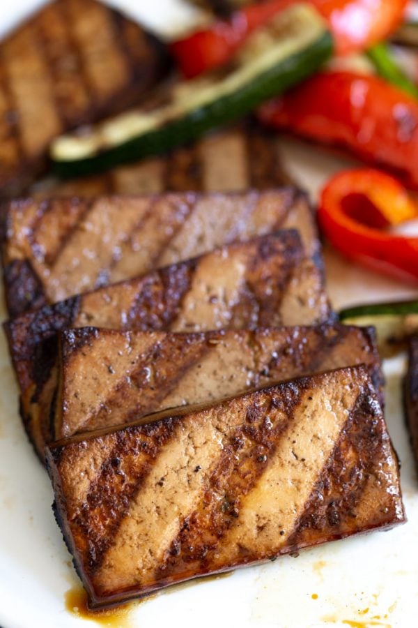 side shot of grilled tofu with grill marks on them. red bell pepper and zucchini in the background