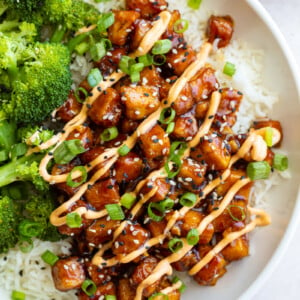 honey garlic tofu in a bowl on top of white rice with cooked broccoli next to it. It's covered in green onion, sesame seeds, and a drizzle of spicy mayo sauce.