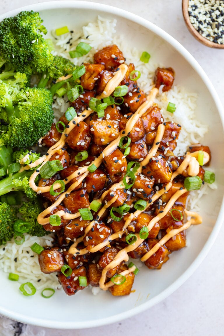 honey garlic tofu in a bowl on top of white rice with cooked broccoli next to it. It's covered in green onion, sesame seeds, and a drizzle of spicy mayo sauce.