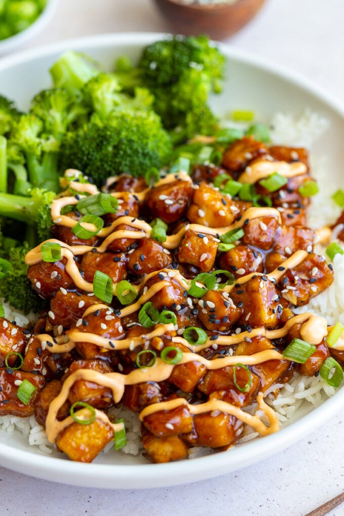 honey garlic tofu in a bowl on top of white rice with cooked broccoli next to it. It's covered in green onion, sesame seeds, and a drizzle of spicy mayo sauce.