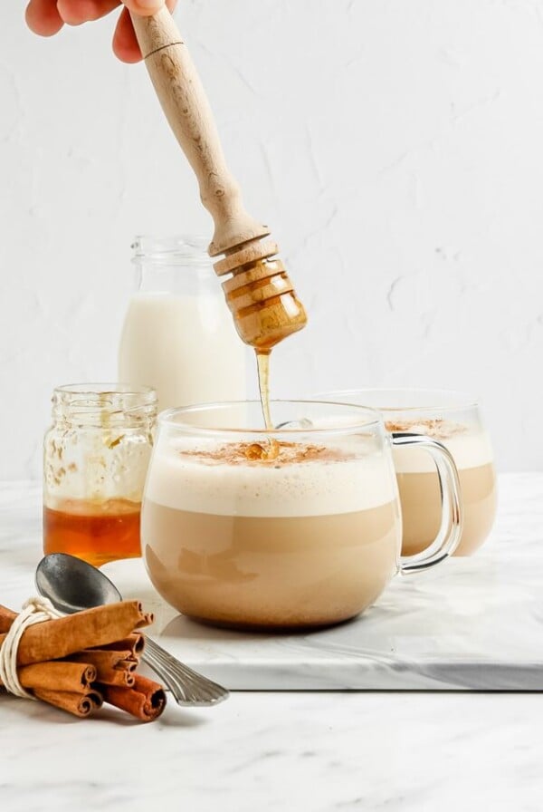 clear mug with a foamed latte in it. a hand drizzling honey on top into the mug