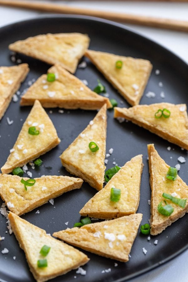 side shot of fried tofu triangles sitting on a white plate and topped with flaky sea salt and green onions