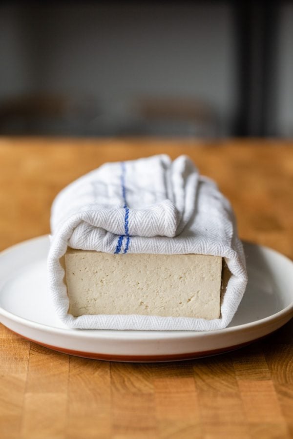 block of tofu wrapped in a white and blue towel, sitting on a white plate. It's all on a butcher block counter