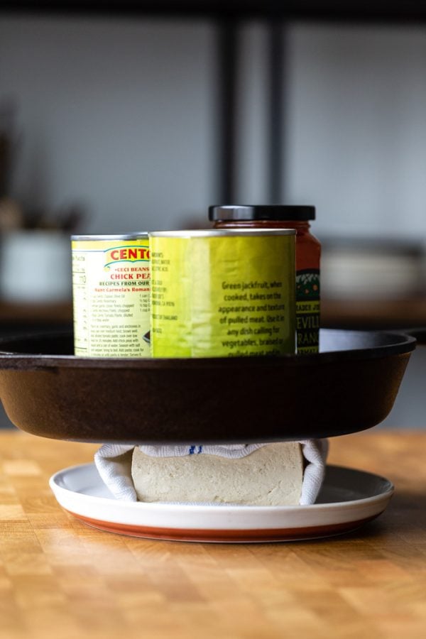 block of tofu wrapped in a white and blue towel, sitting on a white plate. It's all on a butcher block counter. a skillet and heavy cans sitting on top to press the tofu