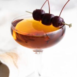 manhattan cocktail (brownish orange liquid) in a coup cocktail glass and garnished with 3 dark maraschino cherries on a gold pick