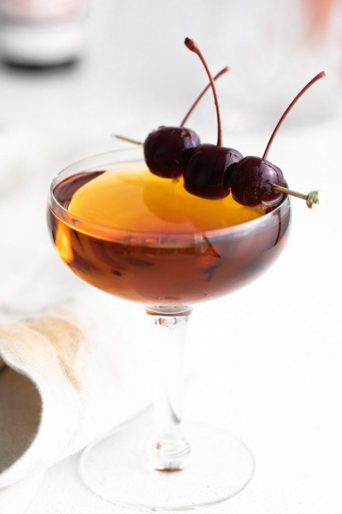 manhattan cocktail (brownish orange liquid) in a coup cocktail glass and garnished with 3 dark maraschino cherries on a gold pick