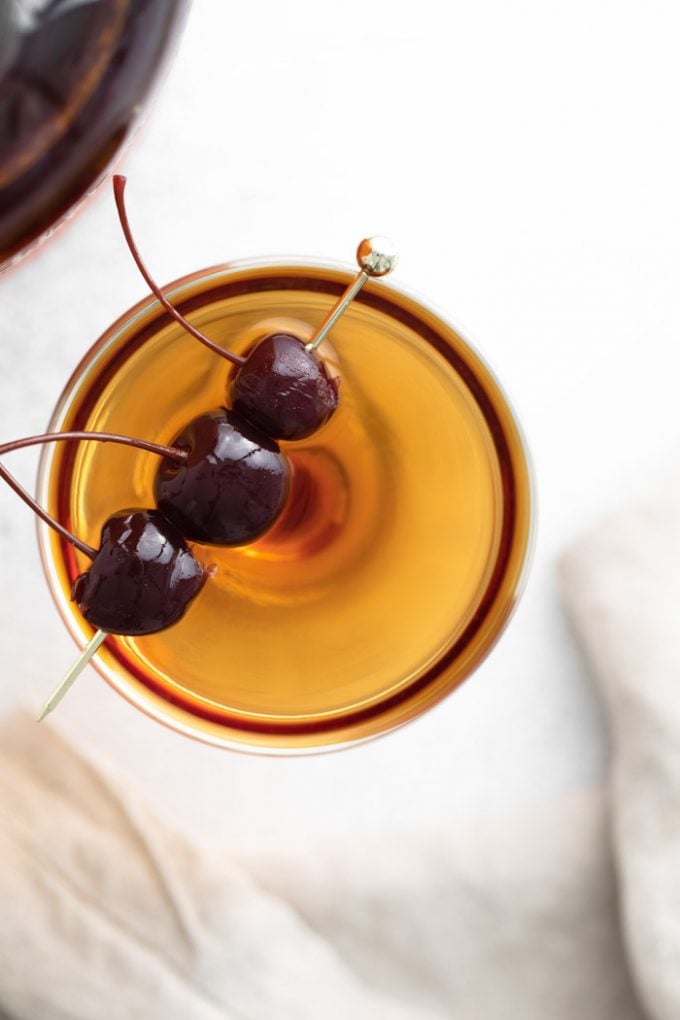 manhattan cocktail (brownish orange liquid) in a coup cocktail glass and garnished with 3 dark maraschino cherries on a gold pick