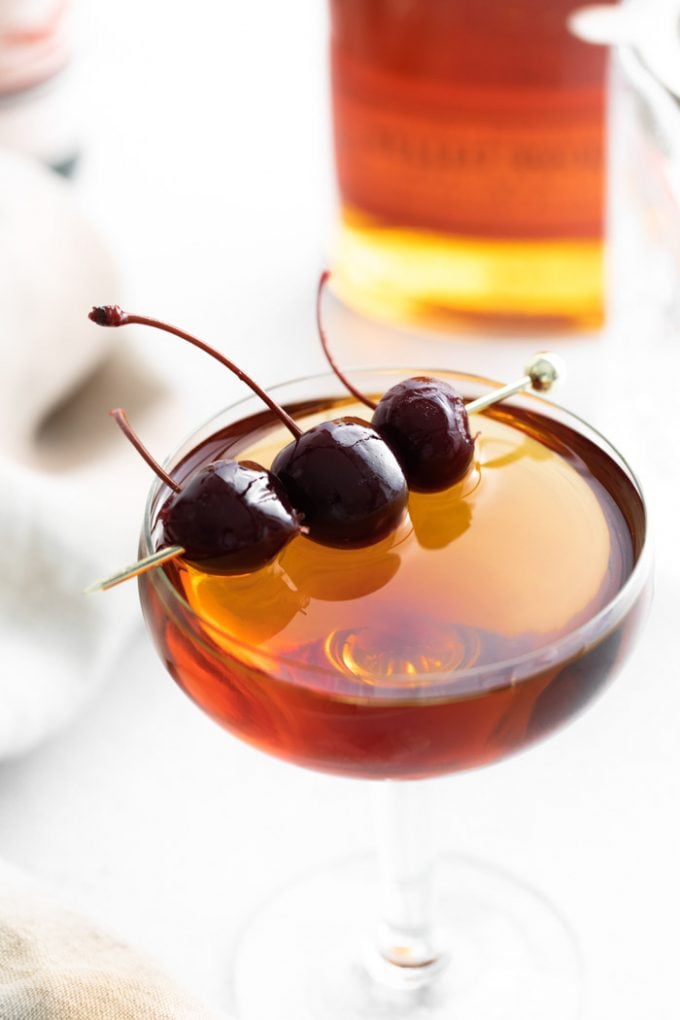 up close shot of manhattan cocktail (brownish orange liquid) in a coup cocktail glass and garnished with 3 dark maraschino cherries on a gold pick