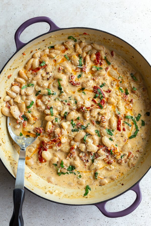 large skillet with creamy butter beans and sundried tomtoes in it known as "marry me butter beans". topped with fresh basil., there's a large spoon on the side