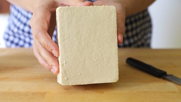 hands holding up a block of firm tofu on top of a butcher clock with a knife in the background