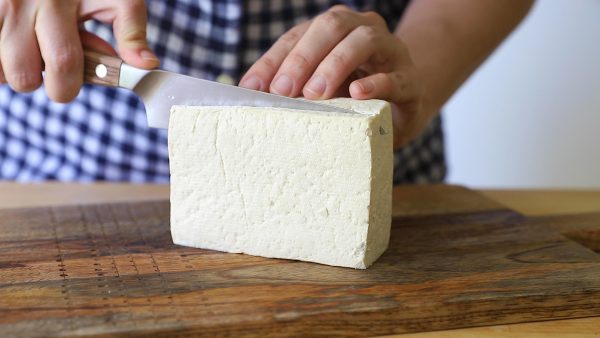 straight on shot of hands holding a block of tofu and cutting it in half on top of a butcher block counter