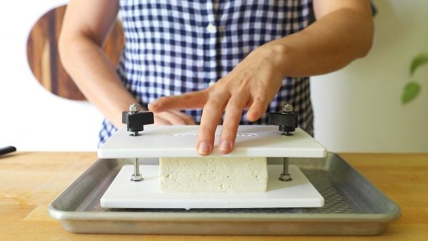 straight on shot of someone placing a black of tofu into a white tofu press that's sitting on top of a baking sheet