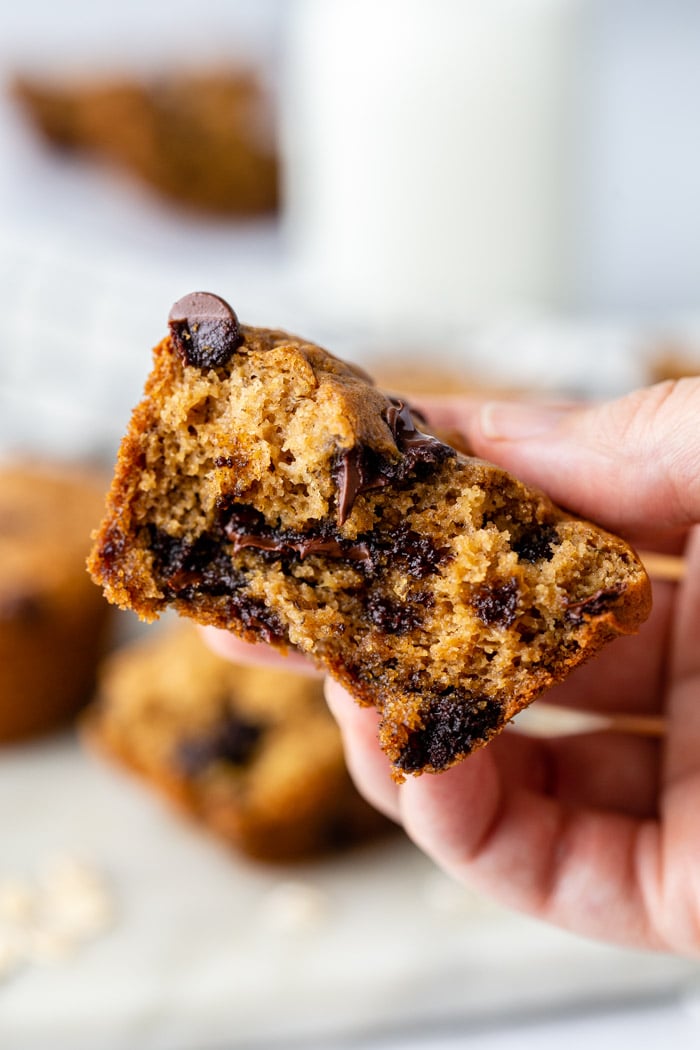 up close shot of a hand holding a half eaten chocolate chip oat flour muffin with more muffins and milk in the background