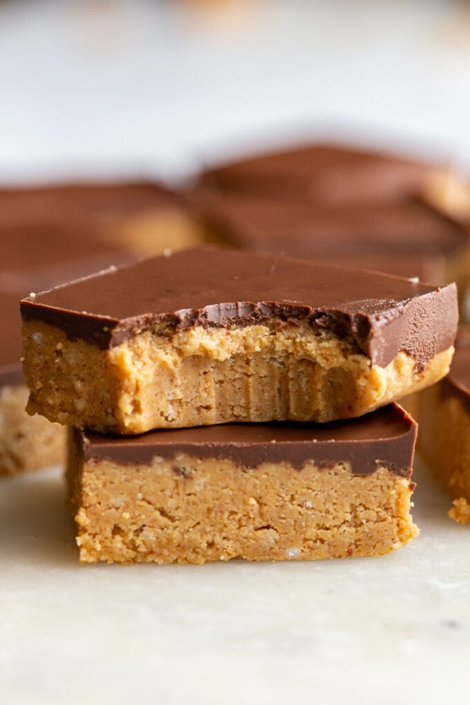 2 chocolate peanut butter bars stacked on top of each other. the top bar has a bite out of it and there are more bars in the background