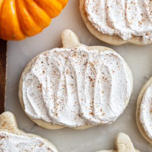 pumpkin shaped sugar cookies topped with a maple icing and sprinkled with nutmeg. a small orange pumpkin in the corner