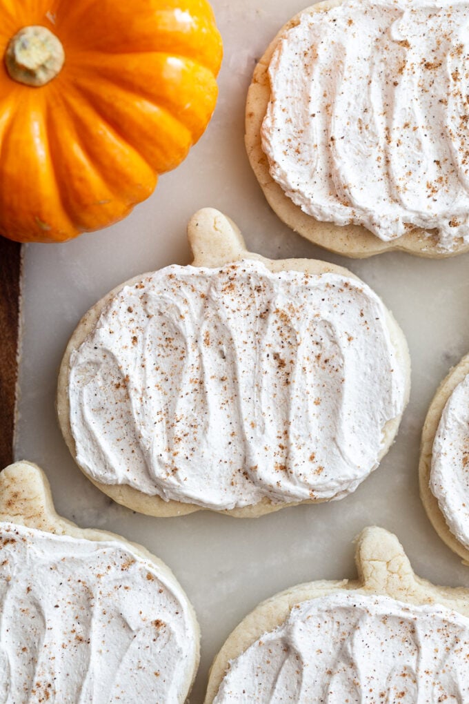 pumpkin shaped sugar cookies topped with a maple icing and sprinkled with nutmeg. a small orange pumpkin in the corner