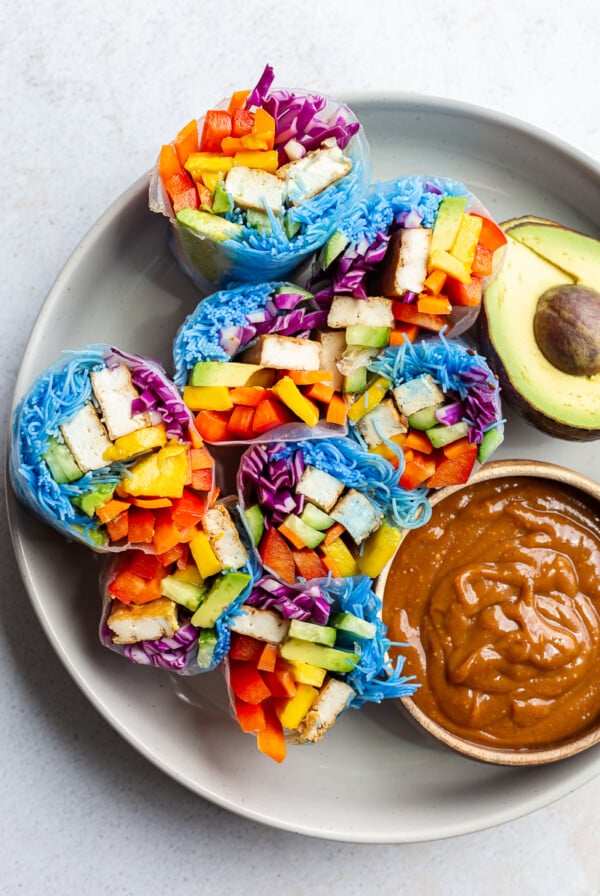 spring rolls made with lots of colorful veggies, and rice noodles that have been dyed blue with blue spirulina. There's also chunks of tofu in them. an avocado and peanut sauce are sitting on the side.