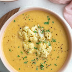 big pink bowl filled with orange cauliflower soup topped with roasted cauliflower and fresh herbs