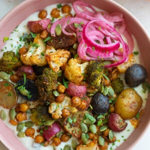 pink bowl filled with roasted veggies including chickpeas, cauliflower, broccoli, and potatoes with tzatziki underneath and pickled onion on the side