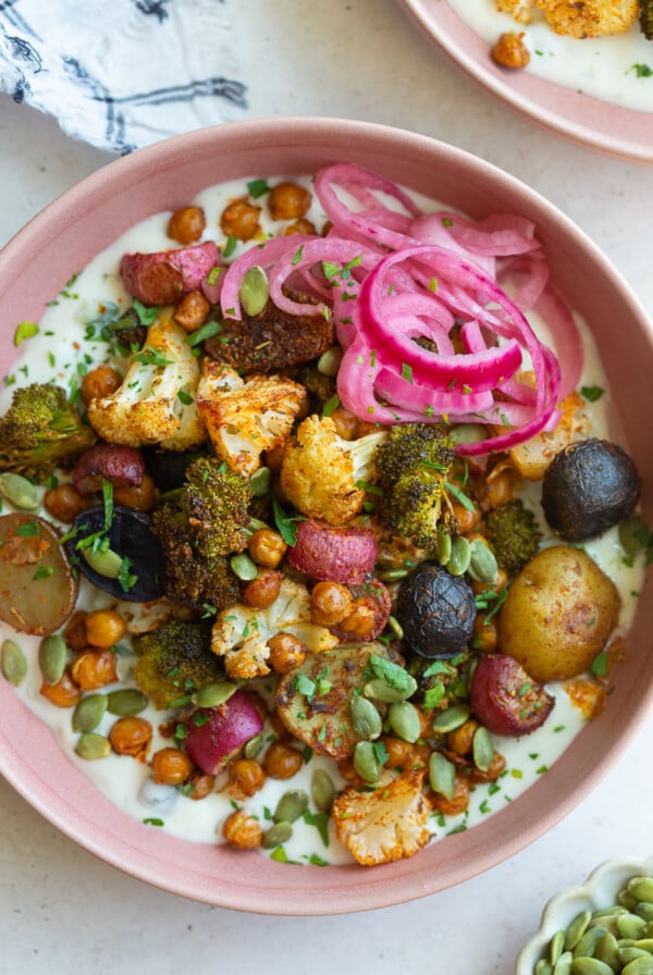 pink bowl filled with roasted veggies including chickpeas, cauliflower, broccoli, and potatoes with tzatziki underneath and pickled onion on the side