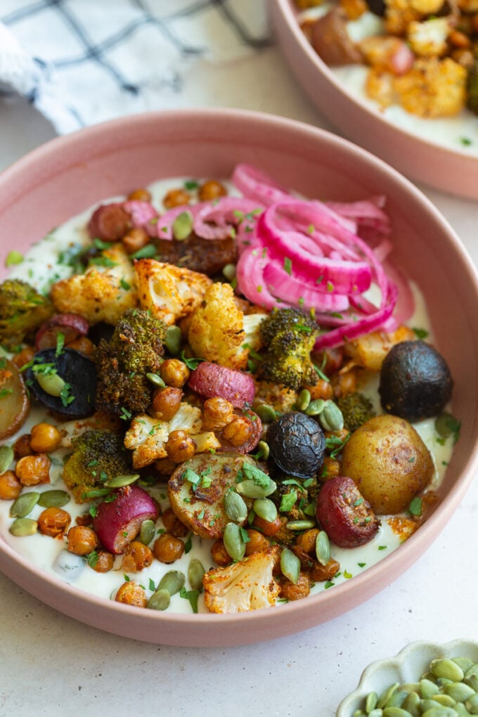 pink bowl filled with roasted veggies including chickpeas, cauliflower, broccoli, and potatoes with tzatziki underneath and pickled onion on the side