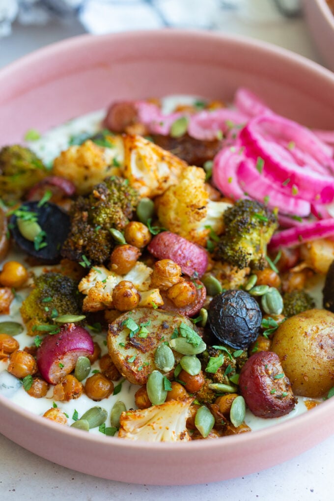 pink bowl filled with roasted veggies including chickpeas, cauliflower, broccoli, and potatoes with tzatziki underneath and pickled onion on the side