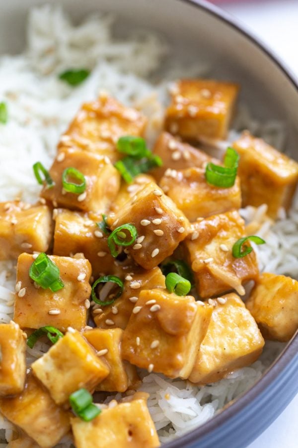 tofu cubes on a bed of white rice and covered in a brown tahini sauce. topped with green onion and sesame seeds