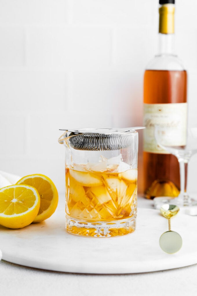 shaker glass with orange liquid and ice inside and cognac bottle in the background