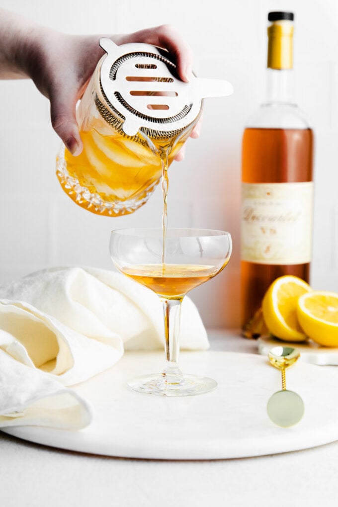 sidecar being poured into a coup glass from a shaker glass with a bottle of cognac in the background