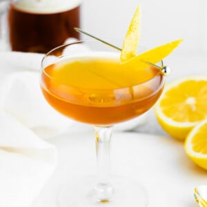 sidecar cocktail in a coupe glass with lemon peel on the side and alcohol bottle on the background as well as oranges