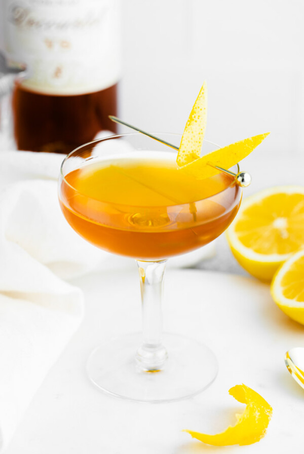 sidecar cocktail in a coupe glass with lemon peel on the side and alcohol bottle on the background as well as oranges