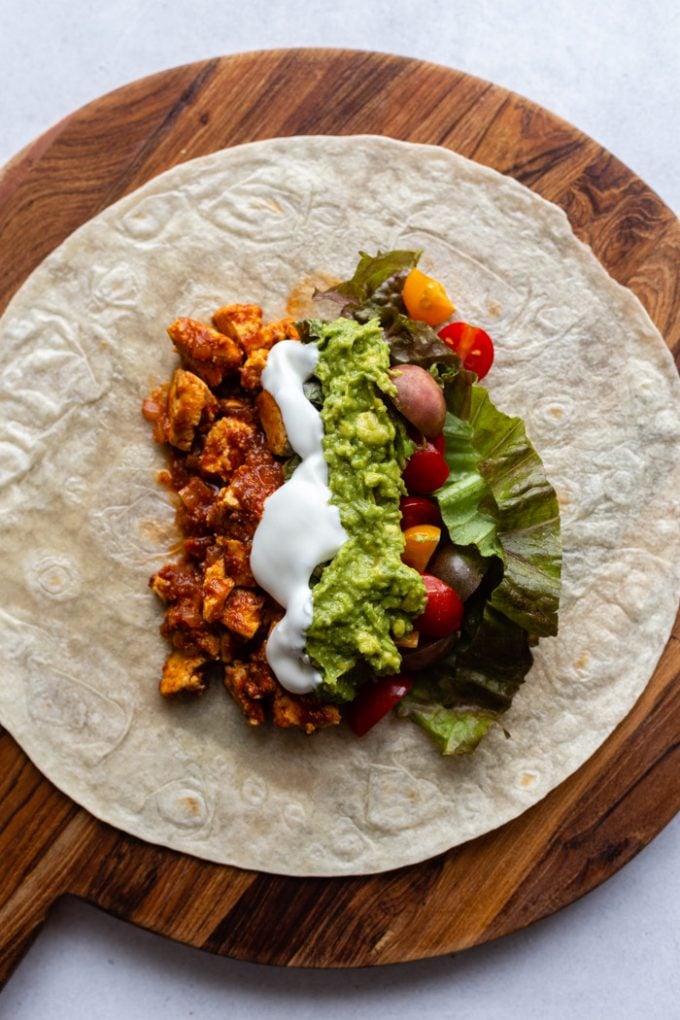 large tortilla sitting on a round wood cutting board. Topped with spicy tofu, sour cream, guacamole, tomatoes, and lettuce