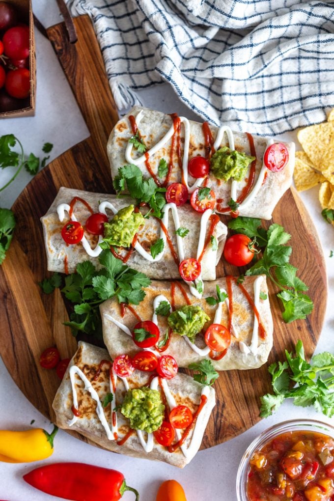 round wood cutting board with 4 grilled burritos on it. the burritos are topped with drizzles of sour cream and hot sauce and sprinkled with cilantro and tomatoes