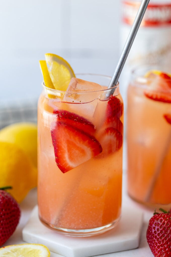 glass filled with strawberry lemonade vodka with a bottle of vodka in the background and garnished with fresh lemon and strawberries