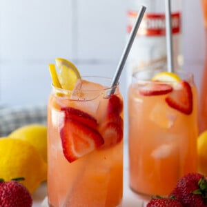 glass filled with strawberry lemonade vodka with a bottle of vodka in the background and garnished with fresh lemon and strawberries