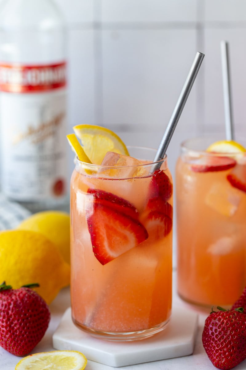 glass filled with strawberry lemonade vodka with a bottle of vodka in the background and garnished with fresh lemon and strawberries