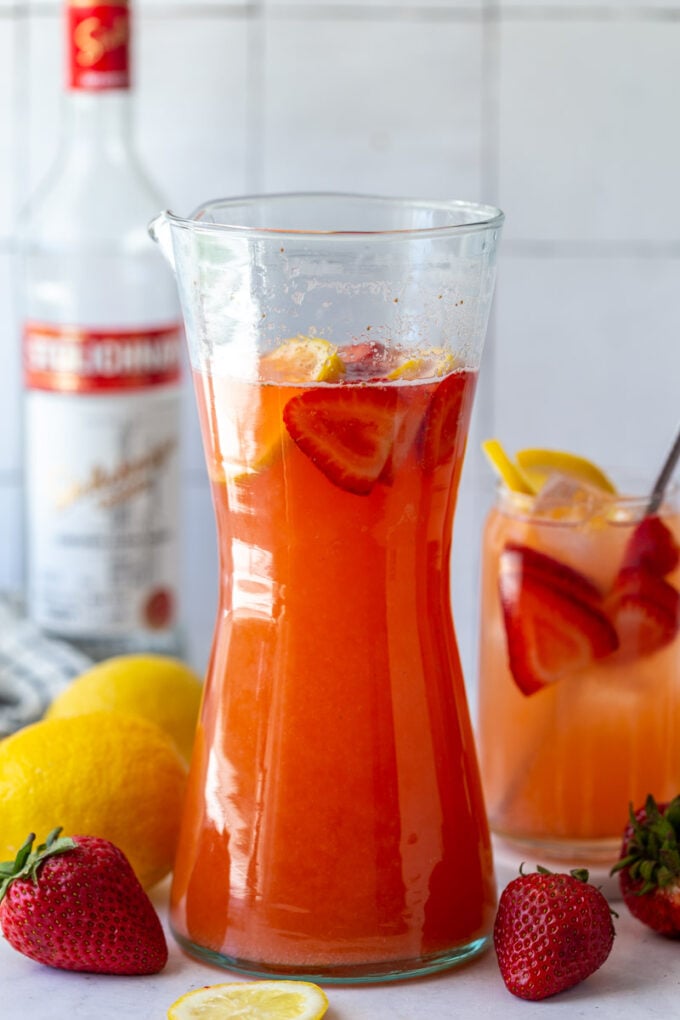 strawberry vodka lemonade in a tall pitcher with fresh fruit around and a bottle of vodka in the background