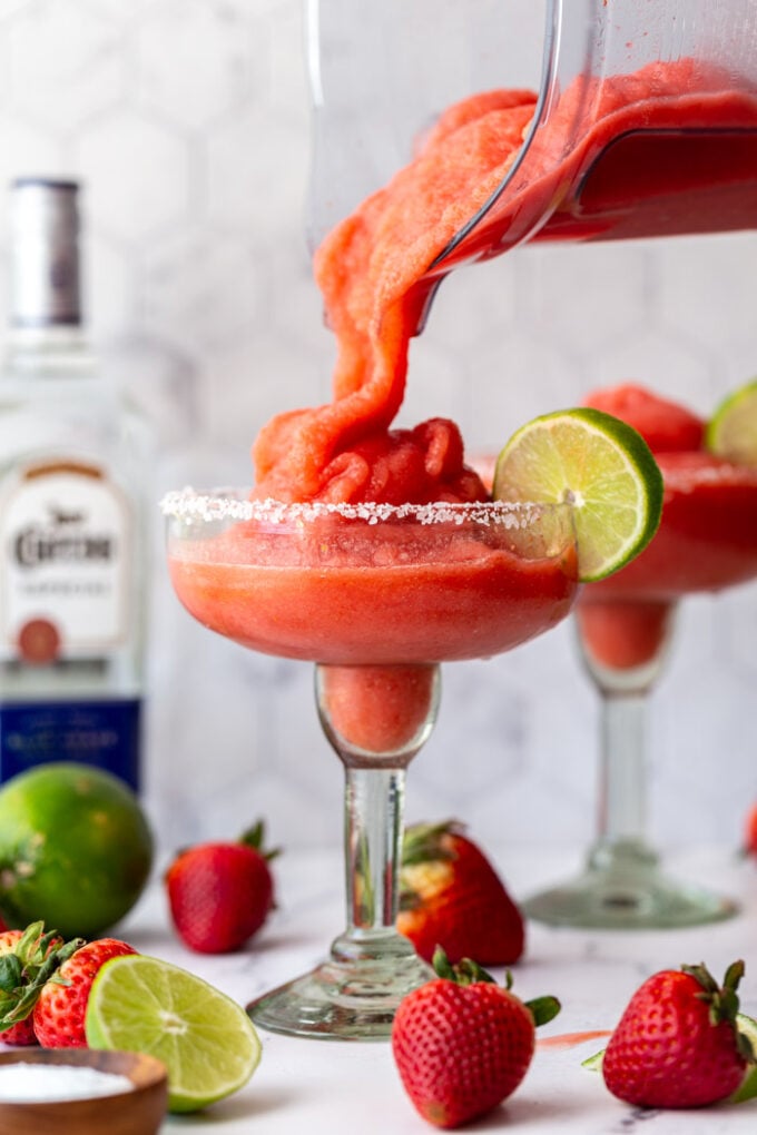 frozen strawberry margarita in a margarita glass being poured from a blender. another drink and a bottle of tequila in the background with fresh fruit around