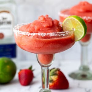 margarita glass filled with a red strawberry frozen margarita and a lime on the rim. more fruit and tequila bottle in the background