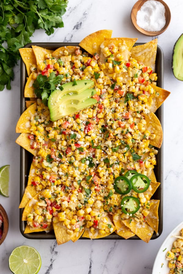 baking tray filled with nachos covered in mexican street cron. cilantro and limes in the corn of the shot