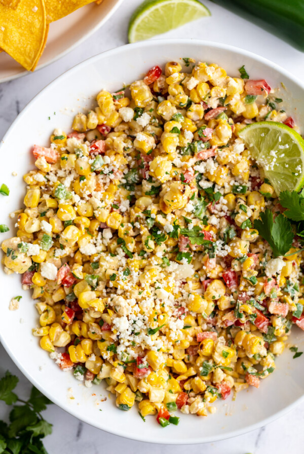 large white bowl of mexican street corn salad topped with cotija cheese and a lime with cilantro in the scene