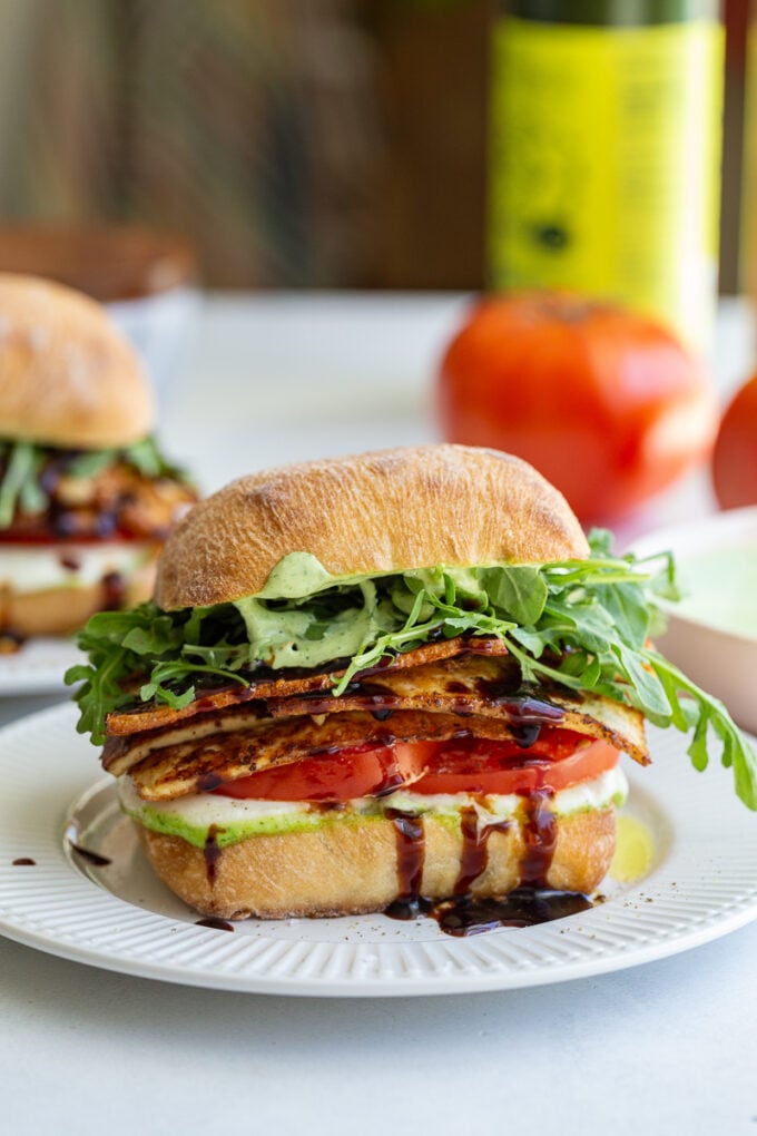 tofu caprese sandwich made with a ciabatta roll and vibrant fresh produce. there's another sandwich and a fresh tomato in the background