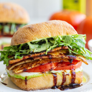 tofu caprese sandwich made with a ciabatta roll and vibrant fresh produce. there's another sandwich and a fresh tomato in the background