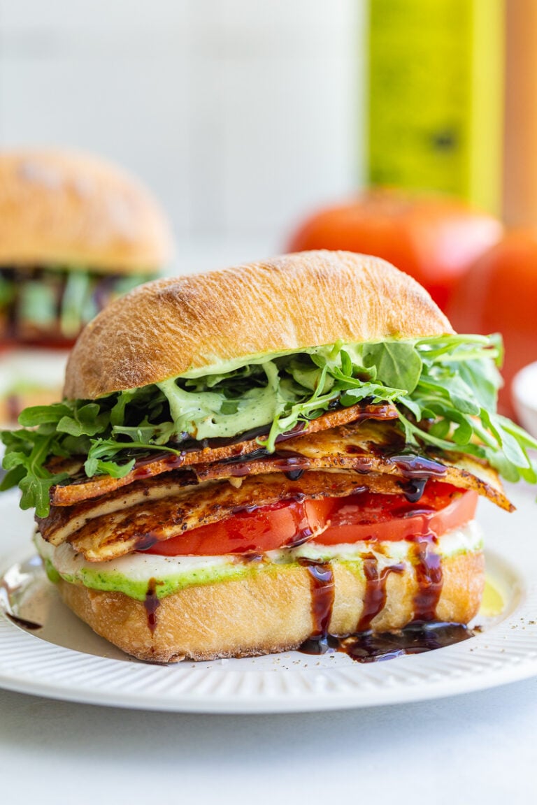 tofu caprese sandwich made with a ciabatta roll and vibrant fresh produce. there's another sandwich and a fresh tomato in the background