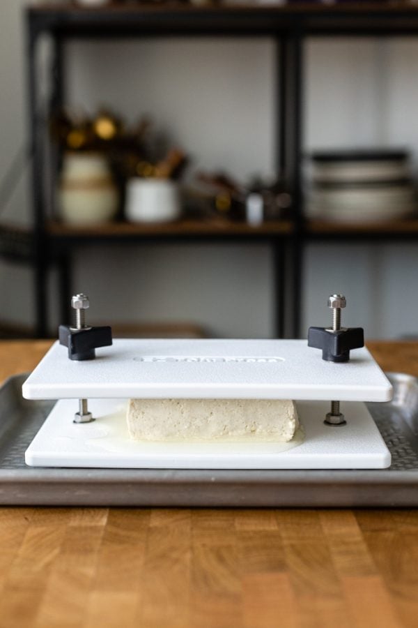 block of tofu in a white tofu press that's in a baking tray sitting on a butcher block counter