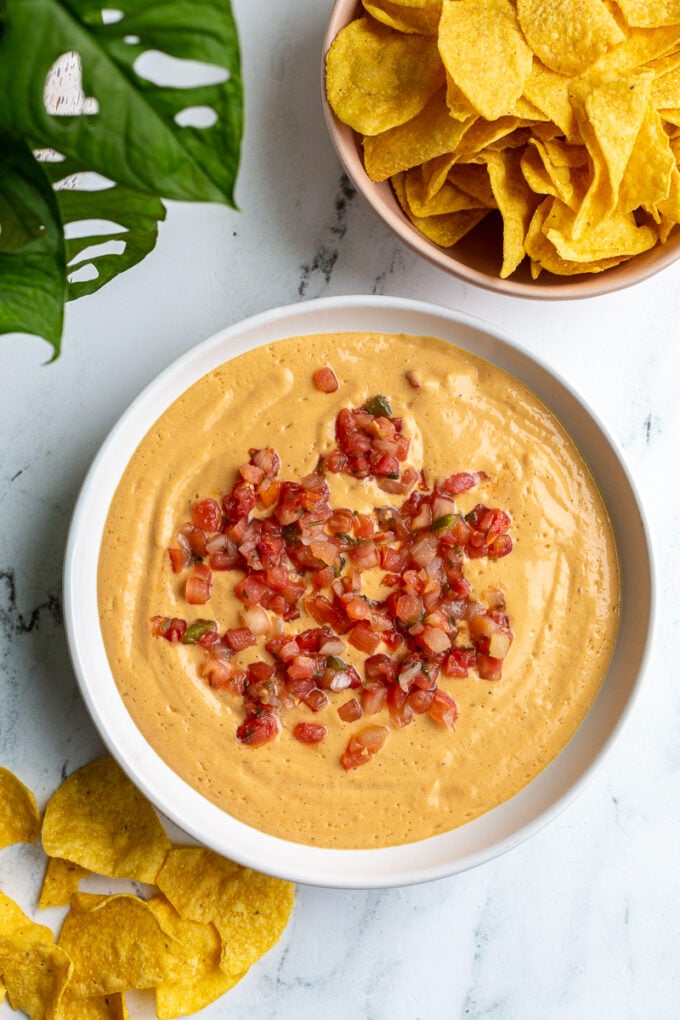 tofu queso cheese dip in a large bowl topped with pico de gallo. there's chips and a plant in the background