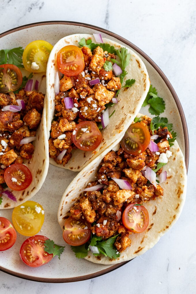 3 flour tortilla filled with tofu taco meat and topped with cherry tomatoes, cilantro, onion, and feta