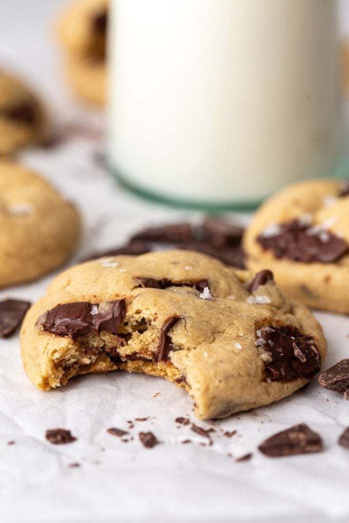 chocolate chip cookie with a bit taken out of it. More cookies, chocolate chips, and glass of milk in the background