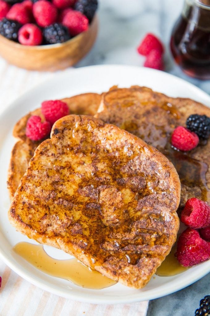 several slices of vegan french toast on a white plate topped with syrup, raspberries and blackberries